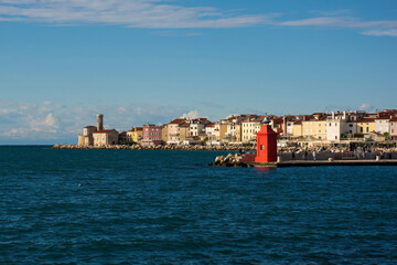 A small red lighthouse on the waterfront of the historic medieval town of Piran on the coast of Slovenia. Background left is the Our Lady of Health Church and a 17th century lighthouse tower
