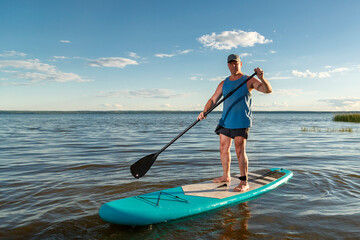 A man in shorts standing on a SUP board with a paddle floats on the water in the rays of the setting sun.