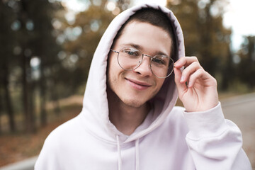 Smiling cute teenager boy 19-20 year old wear glasses and hood over nature background outdoor close...