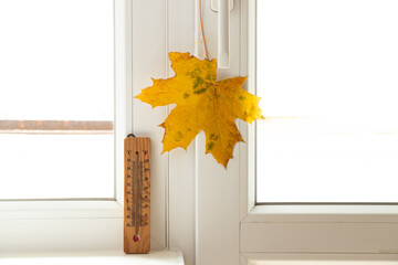 A maple leaf hangs near a thermometer on a window in an apartment, autumn weather outside the window, autumn - obrazy, fototapety, plakaty
