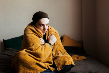 a young man in outerwear is warming himself under a blanket in the bedroom