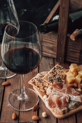 Two glasses of red wine stand on a wooden table. Prosciutto with cheese, red wine, baguette, nuts. Red wine with traditional Mediterranean snacks.