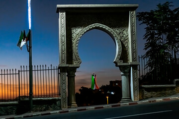 The Martyr's Memorial Monument of Algiers red and green colored lights view through a sculpted door with Ottoman style arch. Flag pole of Algerian flags and a blue sky at twilight golden hour sunlight