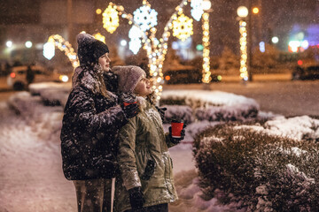 Two teen caucasian sisters wearing warm clothes under falling snow, drinking hot drinks in snowy winter city park decorated with glowing lights for Christmas holidays at night.