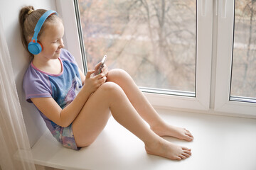 Happy smiling cute girl with smartphone in her hands sits on windowsill and listening music in headphones. Young little girl relaxing at home.