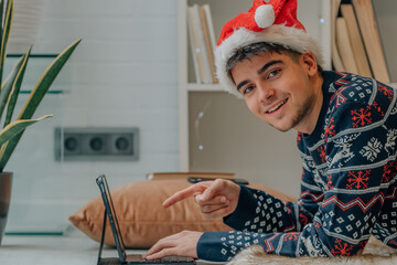 young man pointing laptop screen smiling at home in christmas with santa claus hat