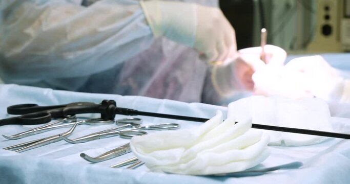 White sterile wipes lie next to the laparoscopic instruments. In the background, the surgeon is suturing the surgical wound with a needle holder, tweezers and needles.