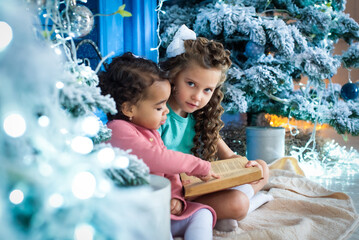 girls with dark and light skin color are reading the book under the christmas tree. african girl