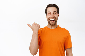 Fototapeta premium Close up of enthusiastic adult man pointing left, smiling amazed, showing advertisement aside, wearing orange t-shirt, white background