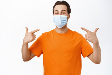 Image of smiling man points at his face with medical face mask, wears orange t-shirt, white background