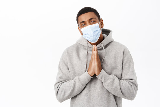 Covid awareness, healthcare concept. Image of concerned african american man in medical mask, begging you, pleading with hopeful look, white background