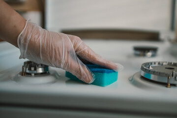 Woman cleaning gas stove with sponge in kitchen, closeup. Closeup of hands wearing rubber gloves while cleaning stove top range with dish sponge
