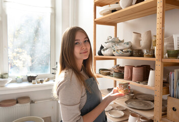 Young beautiful female potter at the ceramic studio. Professional artisan with handmade clay cup.