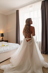 view from the back. a blonde bride wears a white dress by the bedroom window.