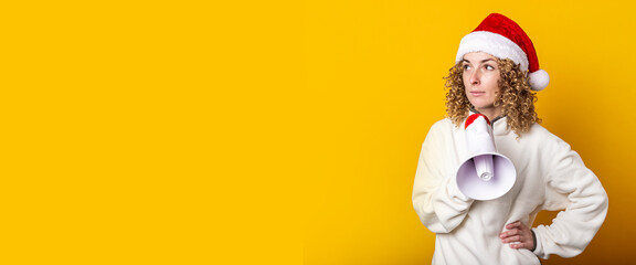 Young woman in santa claus hat with a megaphone on a yellow background. Banner.