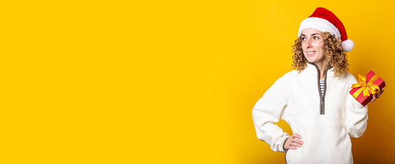 Smiling young woman in santa claus hat holding a gift on a yellow background. Banner.