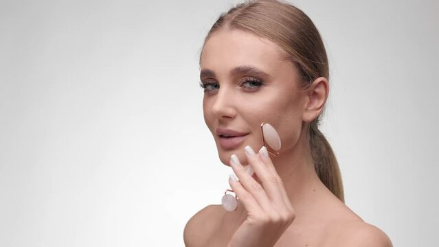 Facial massage. Beautiful blondie woman with healthy fresh skin massaging her face with rose quartz facial roller. Skincare, beauty, anti aging and anti-wrinkle treatment. Cosmetology concept.