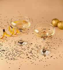 Two glasses filled with champagne with gold sequins and decoration around on the beige background