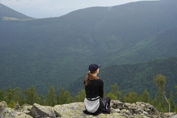 Rear view of young woman in sportswear and cap sitting on rock and watching beautiful view of mountains and forest, horizontal image