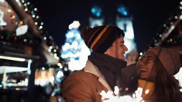 Couple of lovers with sparklers kiss under a Christmas tree on New Year's Day under the chiming of the chimes in the middle of the street in the lights of the Christmas market on the background of the