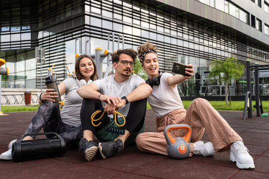 Group of people man and women male and female modern friends before or after training in sitting in open gym modern outdoor fitness park taking selfie photos with mobile phone copy space