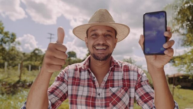 Young man farmer in hat smiling to camera and showing smartphone with chroma key screen. Latin happy male shepherd holding cellphone and demonstrating advertisement. Premium Cinematic 4K