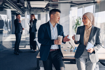 Two happy multiracial colleagues talking and drinking coffee during break time in office