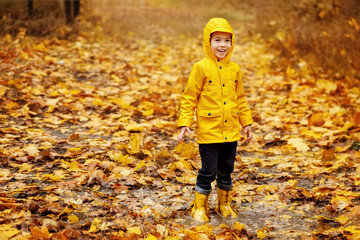 smiling child boy in yellow raincoat and rubber boots stand in puddle in autumn park