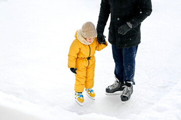 A father holds his little daughter by the hands while standing on the ice rink. Dad teaches the...