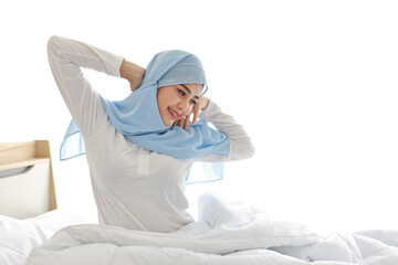 Beautiful asian muslim woman wearing white sleepwear sitting on bed, stretching her arms after...
