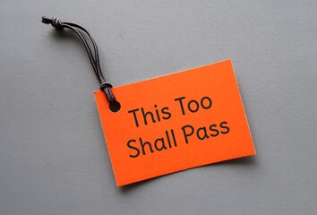 Orange paper tag on gray background with handwritten text THIS TOO SHALL PASS, to remind nothing,...