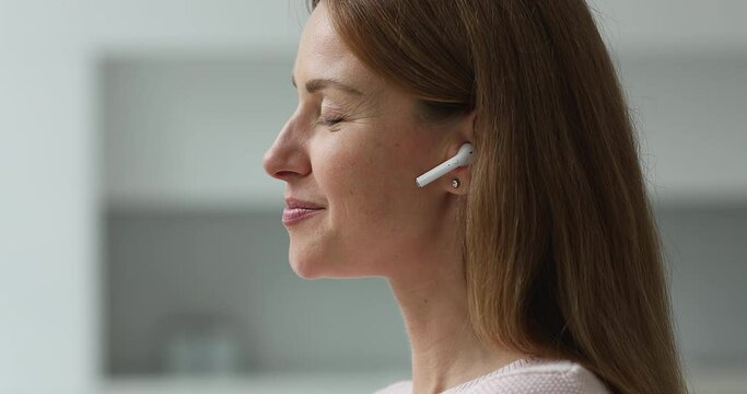 Happy pretty 30s Caucasian woman putting white small wireless earphone into ear, turning player on, listening to music, closing eyes, relaxing with toothy smile, enjoying leisure. Side portrait