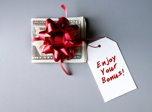 Cash Dollars Money In A Red Ribbon And Gift Bow And White Christmas Gift Tag With Written Text ENJOY YOUR BONUS, Concept Of Bonus Reward At The Year End For Employee