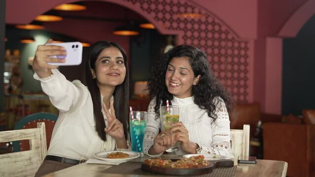 Two Indian business women colleagues Friends eating pizza and smiling for selfie. They are sharing pizza and making selfie photo on smart phone. They are having party drinking juice in the cafe.
