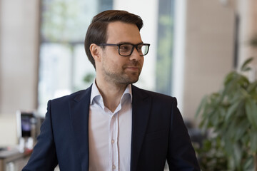 Head shot portrait successful handsome businessman wear glasses and formal suit, looking into distance posing alone standing in modern office workspace. Professional occupation person, workday, career