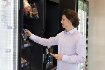 Young businessman or office employee, customer using credit card for payment, paying for snack bought in vending machine at automated retail place using cashless technology. Lunch break, modern tech - obrazy, fototapety, plakaty