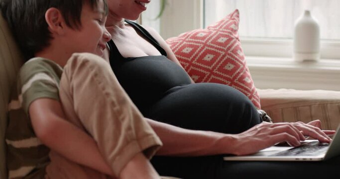 Pregnant mother and 5 year old son sit on couch watching a laptop and laugh - side profile