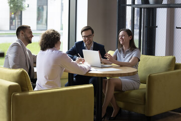 Group of cheerful office employees joking engaged in meeting seated on sofas at table, having...