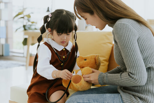 Young asian mother and kid girl daughter with stethoscope sitting on couch role play doctor with bear doll toy together