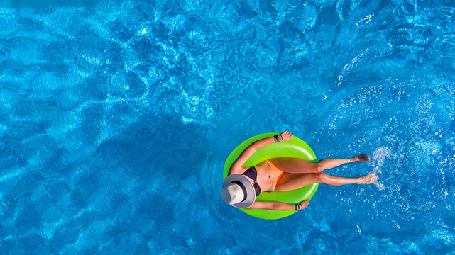 Beautiful woman in hat in swimming pool aerial top view from above, young girl in bikini relaxes and swims on inflatable ring donut and has fun in water on tropical vacation on holiday resort
