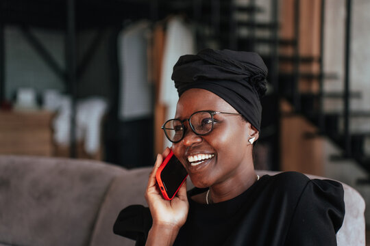 Close up of African young Woman in Black turban, glasses toothy smiling talks by phone sitting at home on cozy sofa. Successful adult African girl speaks with friend using cellphone at home.