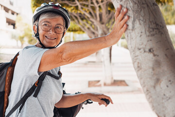 Cheerful cyclist elderly woman in urban park wearing helmet and backpack running with her electro...