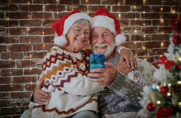 Smiling senior adult couple at home hugging enjoying christmas together at home wearing santa hats. Man and woman video chatting with distant family or friends by phone. Xmas tree