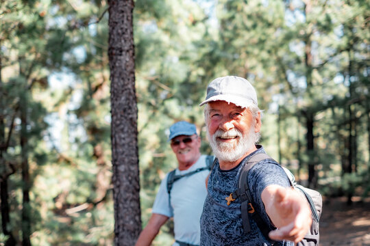 Happy smiling couple of elderly men with hat and backpack look in camera while enjoying mountain hike in the woods appreciating leisure and freedom, retired seniors and healthy lifestyle concept