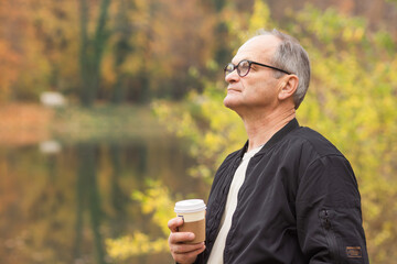 Fototapeta premium Senior man walking and drinking hot coffee in autumn city park. Elderly man holding disposable paper cup in his hands.