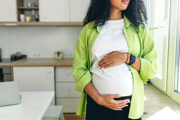 No face picture of pregnant african American woman with long curly hair looking through window standing in kitchen, rubbing her big cute belly, expecting baby boy to be born in few month - 543798018