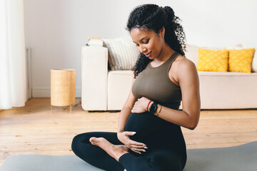 Cute pregnant female of black ethnicity sitting on mat in sports clothes rubbing her adorable...