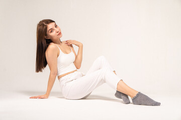 Cute young brown-haired girl in a white top and knitted trousers posing while sitting on a white background