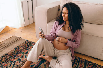 Portrait of gorgeous pregnant female with dark skin and curls sitting on carpet next to couch browsing news feed in her social media app, reading article, watching podcast for future moms - 543798451