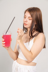 A cute young brown-haired girl drinks a cocktail from a glass with a straw on a white background/The model is dressed in a white top and knitted trousers